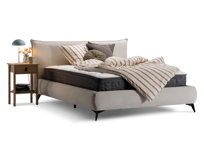 Polstret seng med høj, blød sengegavl i lysebeige stof. Sengen har en tyk, mørkegrå madras og en hvid topmadras. Sengetøjet består af en beige og hvidstribet dyne, flere pyntepuder i forskellige former og neutrale farver samt et matchende pudebetræk. Ved siden af sengen står et natbord i træ med skuffe og hylde, hvorpå der står en blå lampe, en lille vase med blomster og en kop.