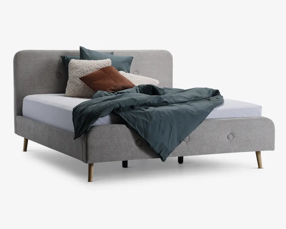 En moderne seng med polstret stel i lysegråt stof, polstret hovedgærde og fodgærde med diskrete knapdetaljer. Sengen står på fire lyse træben. Madrassen er dækket af en mørk petrolfarvet dyne, flere pyntepuder i grønne, brune og cremefarvede nuancer samt en tekstureret cremefarvet pude, hvilket giver sengen et hyggeligt og indbydende udtryk.
