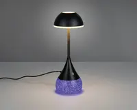 Moderne bordlampe med sort, kuppelformet skærm og slank, lige stang. Basen er konisk med en tekstureret, glitrende lilla overflade, der udsender et blødt lys. Der er en touch-følsom tænd/sluk-knap på den sorte del af basen. Lampen får strøm via en synlig sort ledning.