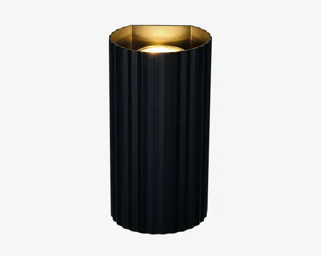 Høj, cylindrisk genstand med en lodret rillet sort yderside og en metallisk gylden inderside. Toppen er åben og afslører en lyskilde, der udsender et varmt skær, som reflekteres af den gyldne belægning for en elegant, stemningsfuld effekt. Designet er moderne og minimalistisk, velegnet til nutidig indretning.
