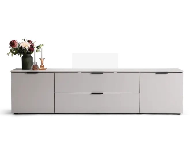 Lav, moderne skænk med mat lysegrå finish, udstyret med to store centrale skuffer og to sidekabinetter med sorte greb. Toppen er dekoreret med en grøn vase med blomster, to hvide lys i lysestager og en lille dekorationsgenstand.