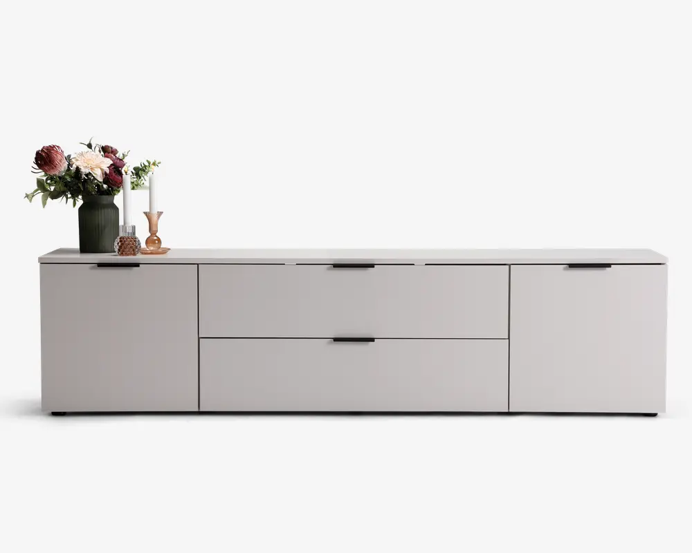 Lav, moderne skænk med mat lysegrå finish, udstyret med to store centrale skuffer og to sidekabinetter med sorte greb. Toppen er dekoreret med en grøn vase med blomster, to hvide lys i lysestager og en lille dekorationsgenstand.