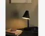 En moderne skrivebordslampe med mat sort finish og en konisk lampeskærm, der leder lyset nedad. Lampen har en slank, lige stander og en rund fod. Den står på et skrivebord sammen med en stak notesbøger, en lille æske, en glaskrukke og en blyant, hvilket skaber en minimalistisk og funktionel arbejdspladsstemning.