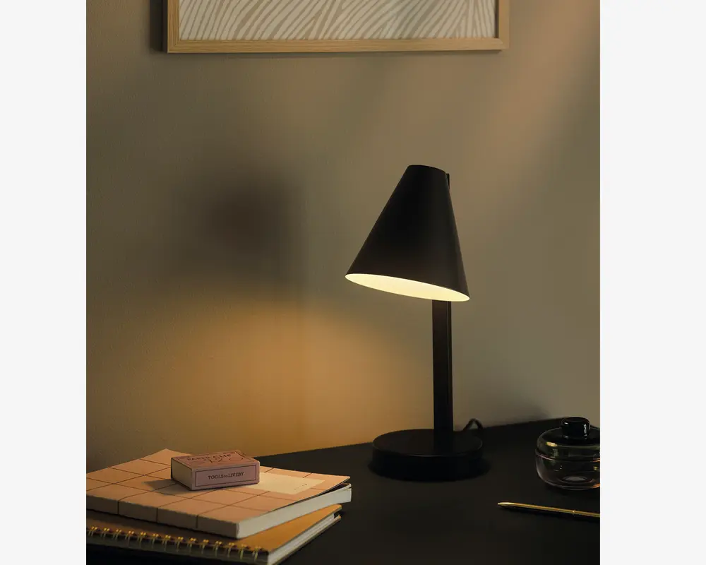 En moderne skrivebordslampe med mat sort finish og en konisk lampeskærm, der leder lyset nedad. Lampen har en slank, lige stander og en rund fod. Den står på et skrivebord sammen med en stak notesbøger, en lille æske, en glaskrukke og en blyant, hvilket skaber en minimalistisk og funktionel arbejdspladsstemning.
