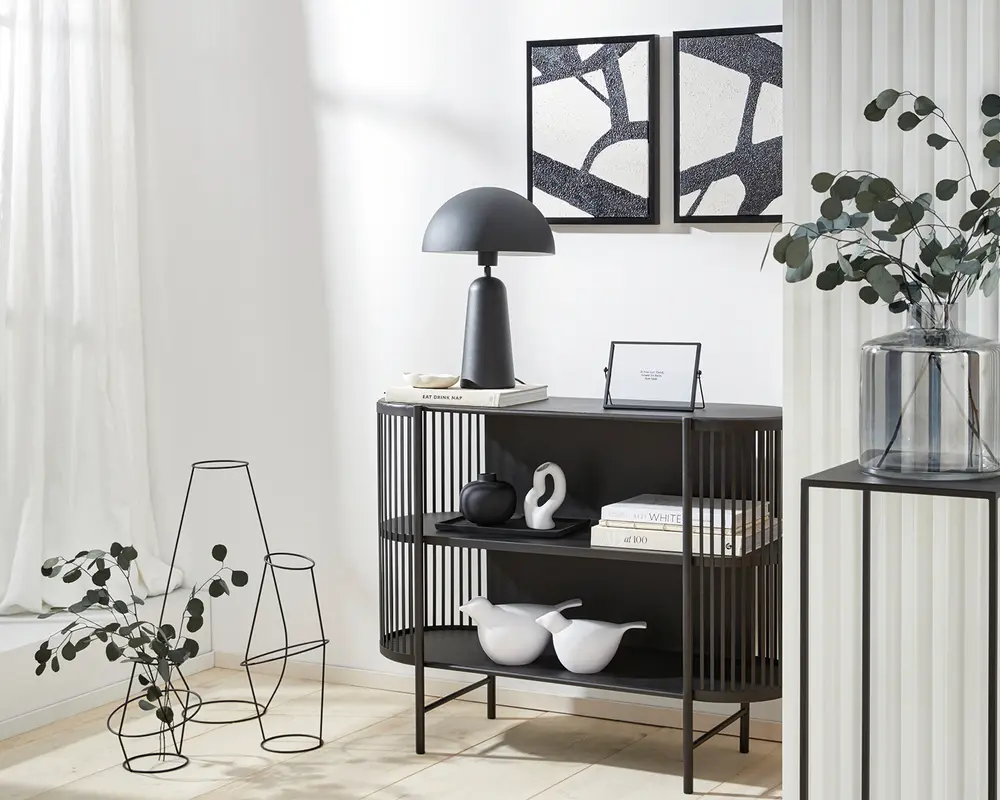 Et moderne, tre-etagers reolsystem med mat sort finish og lodrette lameller på siderne. Hylderne viser dekorative genstande, herunder abstrakte keramikfigurer i sort og hvid, en stak bøger og en indrammet plakat. På øverste hylde står en sort bordlampe med kuppelformet skærm samt en lille bogstak. Reolen står på slanke, lige ben og har et stilrent, minimalistisk design.