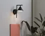 En moderne vægmonteret lampe med mat sort finish, konisk skærm og lige arm, der er fastgjort til en rund base. Lampen udsender et blødt, nedadrettet lys, velegnet til accentbelysning eller læsning. Designet er minimalistisk og nutidigt, og passer til forskellige indretningsstile.