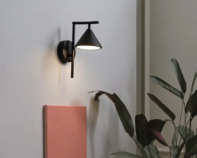 En moderne vægmonteret lampe med mat sort finish, konisk skærm og lige arm, der er fastgjort til en rund base. Lampen udsender et blødt, nedadrettet lys, velegnet til accentbelysning eller læsning. Designet er minimalistisk og nutidigt, og passer til forskellige indretningsstile.