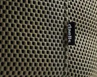 Nærbillede af et khakifarvet, åndbart netstof med bikubemønster. Stoffet har en synlig lodret syning og et lille sort mærke med teksten 'daells' i hvidt, som er fastgjort ved syningen. Netmaterialet ser blødt og fleksibelt ud og er designet til ventilation og komfort.