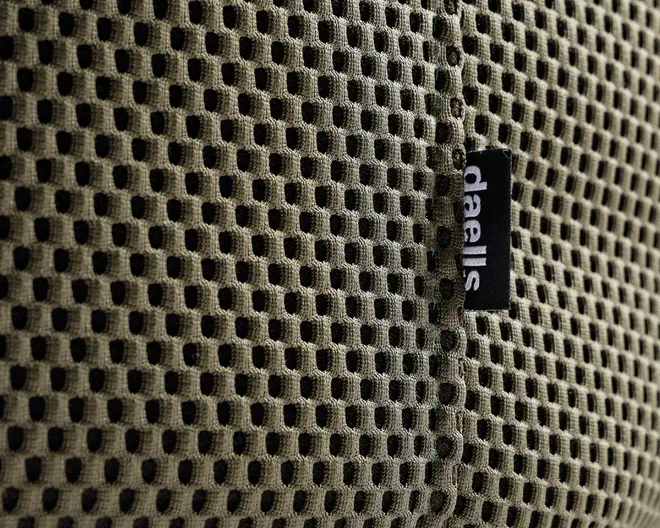 Nærbillede af et khakifarvet, åndbart netstof med bikubemønster. Stoffet har en synlig lodret syning og et lille sort mærke med teksten 'daells' i hvidt, som er fastgjort ved syningen. Netmaterialet ser blødt og fleksibelt ud og er designet til ventilation og komfort.
