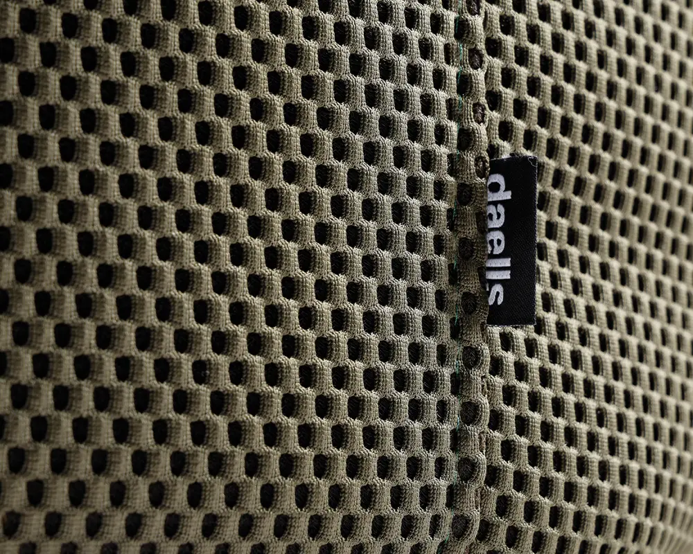 Nærbillede af et khakifarvet, åndbart netstof med bikubemønster. Stoffet har en synlig lodret syning og et lille sort mærke med teksten 'daells' i hvidt, som er fastgjort ved syningen. Netmaterialet ser blødt og fleksibelt ud og er designet til ventilation og komfort.