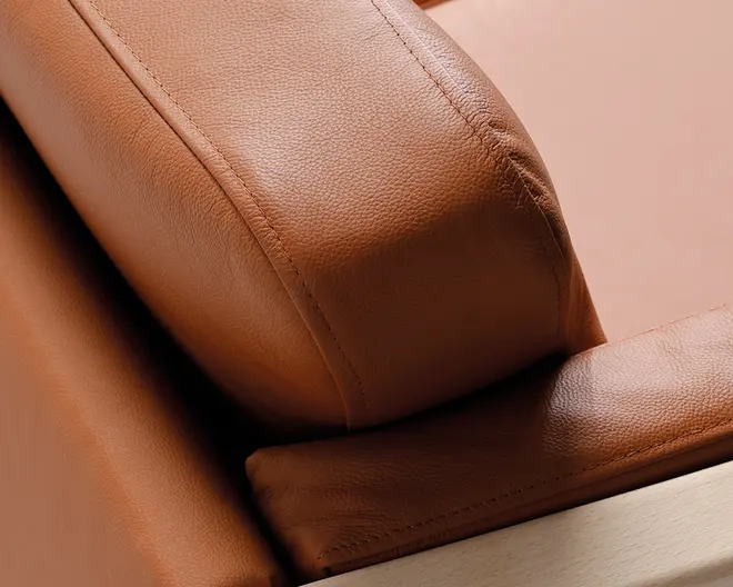 Nærbillede af et møbel betrukket med glat, lysebrunt læder med synlige syninger langs kanterne. Læderet har en let nubret struktur, og hynden ser tyk og velpolstret ud. Møblets bund er lavet af lyst træ, hvilket giver en moderne og elegant kontrast til læderet.