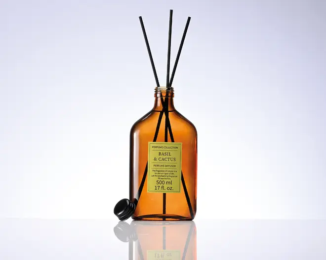 Ravfarvet glasflaske til duftpinde med gul etiket med teksten "Basil & Cactus", tre sorte duftpinde og en sort kapsel lagt ved siden af på en reflekterende overflade.