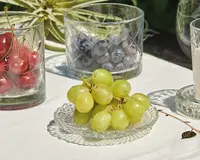 En lille, klar glas-tallerken med en klase friske grønne druer, der har glat, blankt skind og en rund, fyldig form. Tallerkenen har et mønster med små prikker. I baggrunden står to klare glas med diamantmønster, det ene fyldt med røde kirsebær og det andet med blåbær. Produkterne er anrettet på en hvid dug, hvilket giver et rent og lyst udtryk.