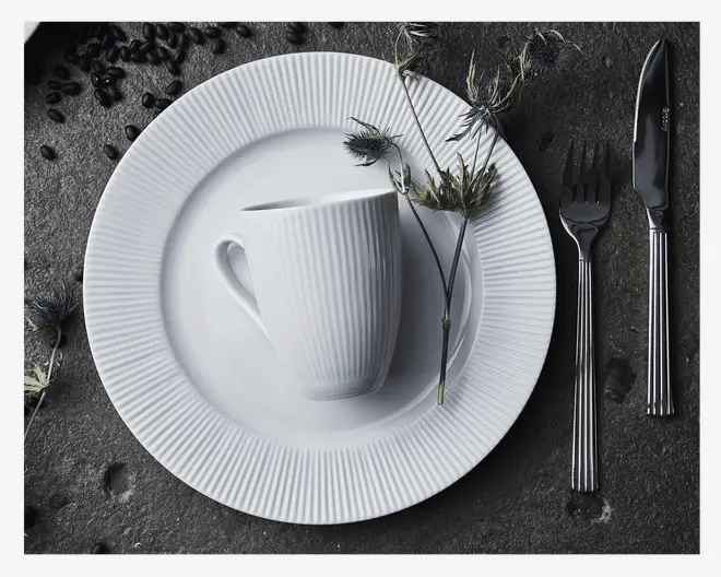 Et hvidt keramik krus og tallerkensæt med en diskret lodret rillet tekstur. Kruset står på en matchende underkop, som er placeret på en større middagstallerken, alle med samme rillede design. Sættet ledsages af en gaffel og kniv i rustfrit stål med stribede håndtag. Dekorative tørrede blomster og spredte kaffebønner er arrangeret omkring sættet på en mørk stens overflade.