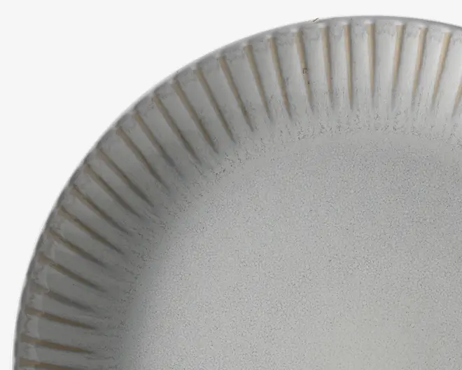 Nærbillede af en rund tallerken med mat, lysegrå og let plettet overflade. Kanten har et hævet, lodret rillet mønster, som giver tallerkenen en tekstureret og elegant fremtoning. Farven skifter diskret fra kanten mod midten og tilføjer dybde til designet.
