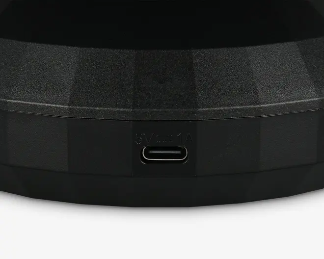 Nærbillede af en sort, tekstureret overflade med en USB-C opladningsport. Over porten er teksten '5V⎓1A' indgraveret, hvilket angiver strømindgangens specifikationer. Overfladen har et facetformet, geometrisk design.