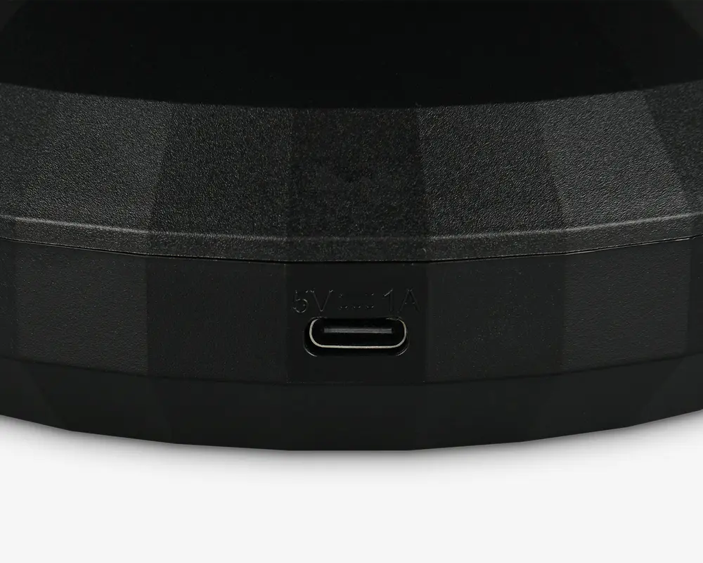 Nærbillede af en sort, tekstureret overflade med en USB-C opladningsport. Over porten er teksten '5V⎓1A' indgraveret, hvilket angiver strømindgangens specifikationer. Overfladen har et facetformet, geometrisk design.