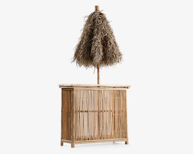 Fritstående bardisk fremstillet af naturlig bambus med lodrette lameller på front og sider. Baren har en matchende bambusbordplade og er udstyret med en stor, dekorativ parasol lavet af tørret græs eller palmeblade, hvilket giver et tropisk, tiki-inspireret udseende. Det samlede design har et rustikt, håndlavet præg, der egner sig til udendørs eller strandtema-miljøer.