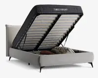 Polstret senge ramme med løftbar lamelbund, der afslører et rummeligt opbevaringsrum under sengen. Opbevaringsområdet indeholder puder og sengetøj. Sengen har et lyst stofbetræk og sorte ben, og madrassens støtte er hævet for nem adgang.