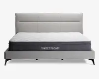 En moderne seng med et lysegråt polstret hovedgærde og ramme, udstyret med en tyk madras med quiltet hvid top og mørkegrå sider. Madrassen har mærket 'SWEETNIGHT' broderet på siden. Hovedgærdet har et stilrent, panelopdelt design, og sengen står på korte, skråtstillede sorte ben.