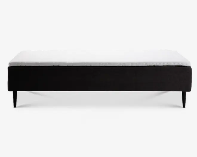 Rektangulær sengebund med et minimalistisk design, der har en sort stofbeklædt ramme og slanke, tilspidsede ben. Den øverste flade er dækket af en hvid madras eller topmadras, hvilket giver et rent og moderne udtryk. Sengebunden er hævet fra gulvet, hvilket gør det nemt at rengøre under.