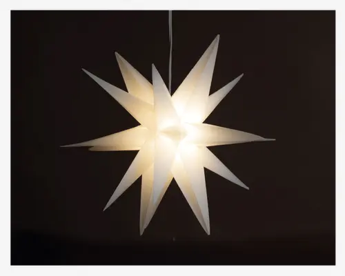 Hængende lampe med tredimensionel stjerneform, der har flere aflange spidser, som stråler ud fra midten. Materialet virker gennemsigtigt og lader et blødt, varmt lys skinne igennem. Designet er moderne og dekorativt, velegnet til festlig eller stemningsfuld belysning.