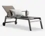 En moderne udendørs liggestol med en stilren, vinklet metalramme i mat finish. Stolen har et justerbart ryglæn og sæde i taupefarvet, slidstærkt netstof. Den er udstyret med to solide armlæn og hjul på bagbenene for nem flytning. Liggestolen er pyntet med en stribet pude for ekstra komfort, et sammenrullet magasin, en dekorativ bakke og en farverig termokop med sugerør placeret på sædet.