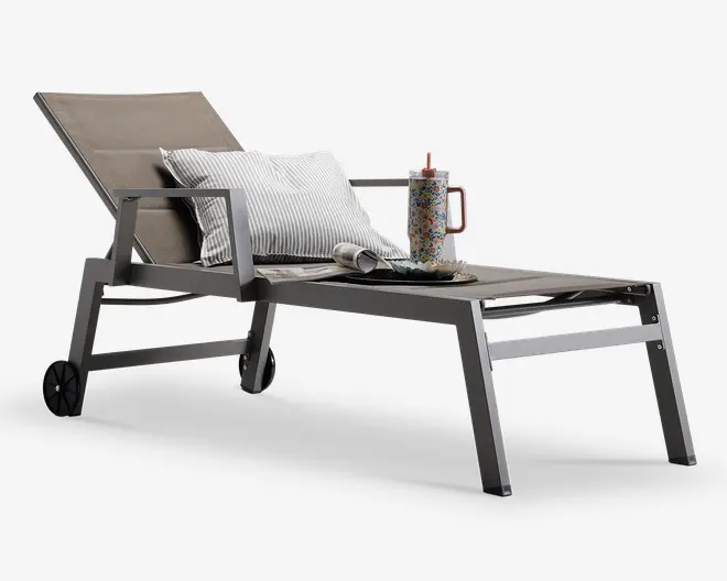 En moderne udendørs liggestol med en stilren, vinklet metalramme i mat finish. Stolen har et justerbart ryglæn og sæde i taupefarvet, slidstærkt netstof. Den er udstyret med to solide armlæn og hjul på bagbenene for nem flytning. Liggestolen er pyntet med en stribet pude for ekstra komfort, et sammenrullet magasin, en dekorativ bakke og en farverig termokop med sugerør placeret på sædet.