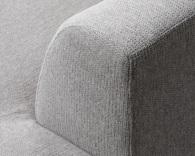 Nærbillede af et sofaarmlæn betrukket med lysegråt, tekstureret stof. Armlænet har en moderne, firkantet form med bløde, afrundede kanter, og stoffet ser tæt vævet ud, hvilket giver et holdbart og behageligt udtryk.