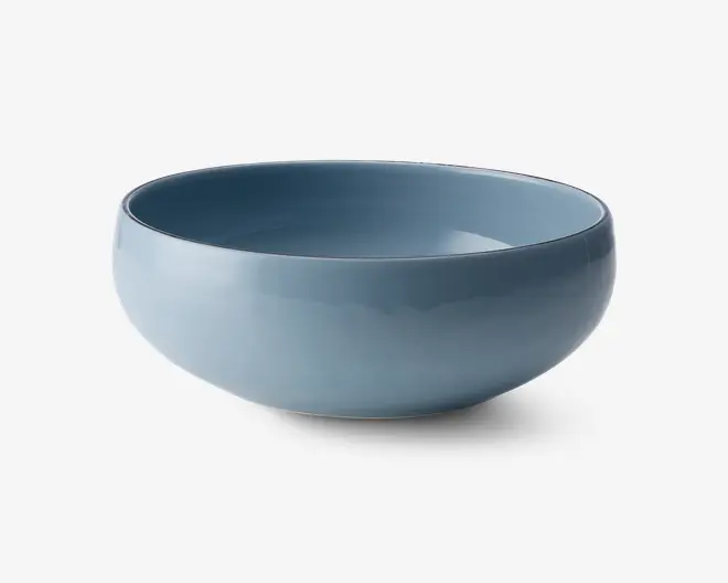 Rund skål med en glat, blank overflade i en afdæmpet blå farve. Skålen har et enkelt, moderne design med blødt buede sider og en let tyk kant. Både indersiden og ydersiden er ensfarvet, og skålen ser ud til at være lavet af keramik eller porcelæn.