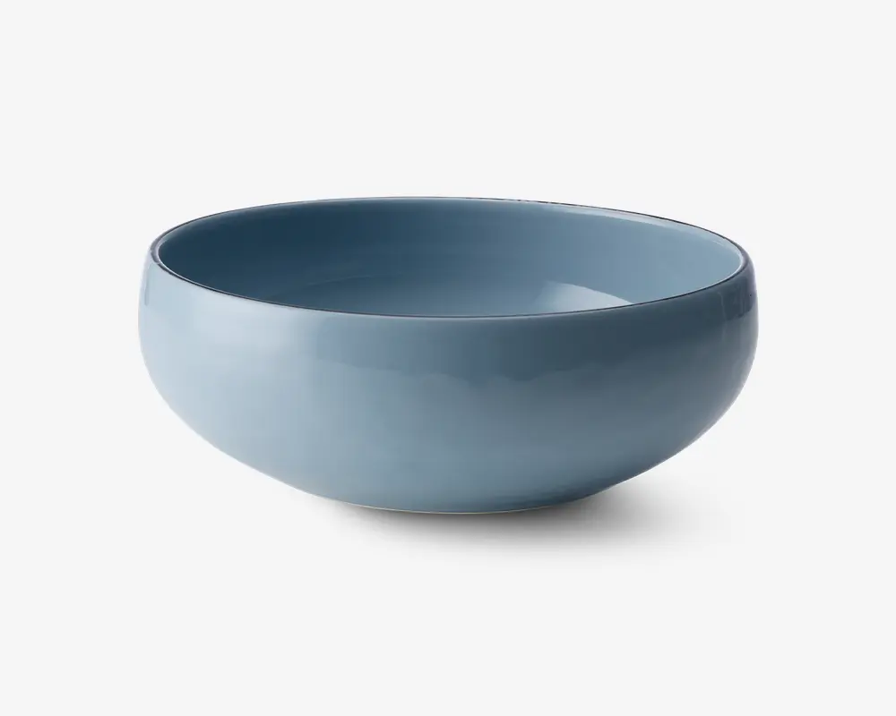 Rund skål med en glat, blank overflade i en afdæmpet blå farve. Skålen har et enkelt, moderne design med blødt buede sider og en let tyk kant. Både indersiden og ydersiden er ensfarvet, og skålen ser ud til at være lavet af keramik eller porcelæn.