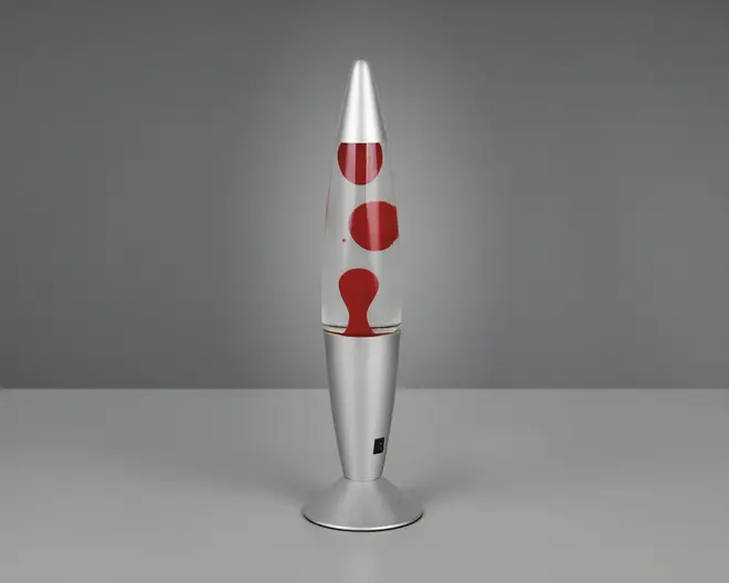 En dekorativ lampe med en slank, metallisk sølvfarvet base og top, der har en klar glasbeholder fyldt med røde voksbobler i væske. Lampen har en moderne, raketformet udseende, og voksen danner organiske, afrundede former indeni, når den opvarmes. Der er en lille kontakt på basen til betjening.