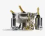 Et festligt arrangement med en stor, riflede isspand i rustfrit stål, der indeholder tre flasker champagne med gyldne foliepropper. Ved siden af spanden står en matchende cocktailshaker i rustfrit stål og en høj, cylindrisk vinkøler med endnu en flaske. Scenen er pyntet med små sølvfarvede diskokugler og guld- og hvidt krøllet bånd, hvilket skaber en festlig stemning.