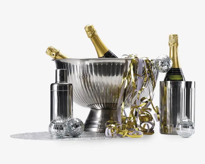 Et festligt arrangement med en stor, riflede isspand i rustfrit stål, der indeholder tre flasker champagne med gyldne foliepropper. Ved siden af spanden står en matchende cocktailshaker i rustfrit stål og en høj, cylindrisk vinkøler med endnu en flaske. Scenen er pyntet med små sølvfarvede diskokugler og guld- og hvidt krøllet bånd, hvilket skaber en festlig stemning.