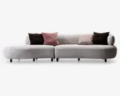 Moderne sofa med sektioner, betrukket med lysegråt, blødt stof. Designet har afrundede kanter og lav ryg, samt en chaiselong til venstre. Sofaen er pyntet med fem pyntepuder: to i mørkebrun, én i ensfarvet rosa, én i rosa med geometrisk mønster og én i beige med cirkelmønster. Sofaen står på korte, sorte ben.