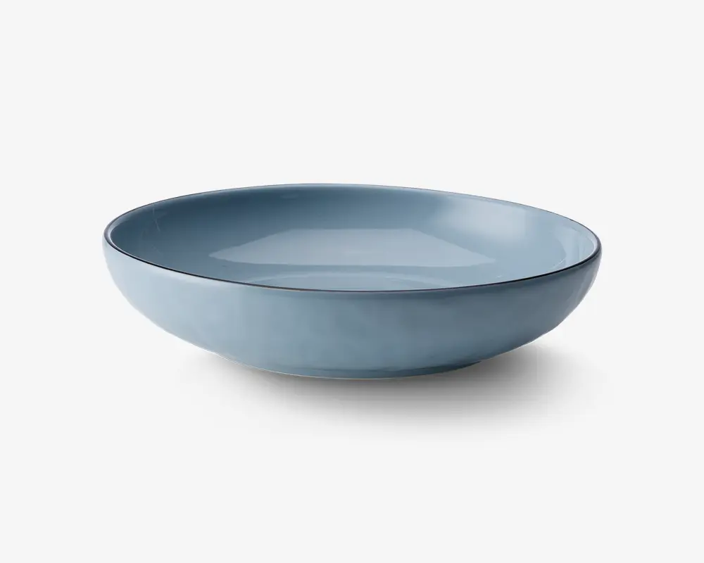 En rund, lav skål med en glat mat blå yderside og en blank blå inderside. Skålens kant er let hævet og har en diskret mørk kant, hvilket giver skålen et moderne og elegant udtryk.