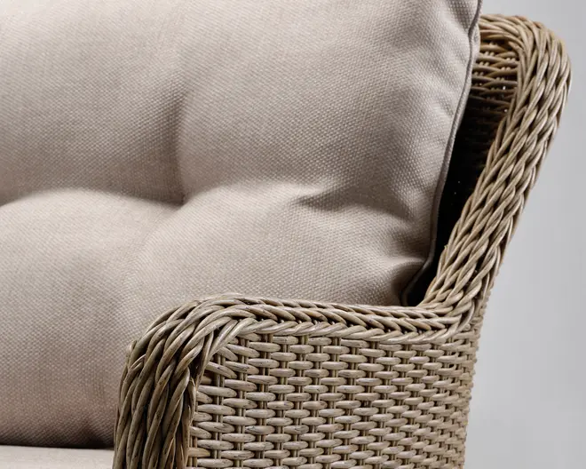 Nærbillede af en stol med tæt flettet rattan- eller kurveramme i en naturlig beige farve. Stolen har tykke, polstrede hynder betrukket med et lyst beige stof, som giver en blød og komfortabel siddeoplevelse. Armlænet er glat buet og fremhæver det detaljerede fletmønster.