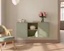 Vægmonteret skab i en afdæmpet salviegrøn farve med et minimalistisk design. Skabet har to lukkede rum med enkle, grebsfri låger og to åbne hylder i midten. De åbne hylder viser en dekorativ fletkurv, en lille stak bøger og et skulpturelt objekt. Ovenpå skabet står en glasvase med lyserøde blomster, en stak bøger og et sæt keramikskåle. Udtrykket er rent og moderne med blødt, naturligt lys og lyst trægulv.