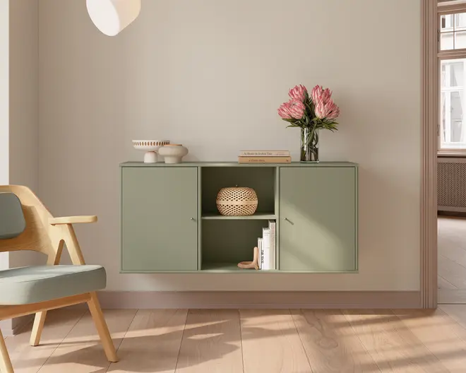 Vægmonteret skab i en afdæmpet salviegrøn farve med et minimalistisk design. Skabet har to lukkede rum med enkle, grebsfri låger og to åbne hylder i midten. De åbne hylder viser en dekorativ fletkurv, en lille stak bøger og et skulpturelt objekt. Ovenpå skabet står en glasvase med lyserøde blomster, en stak bøger og et sæt keramikskåle. Udtrykket er rent og moderne med blødt, naturligt lys og lyst trægulv.