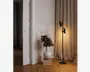 En moderne sort gulvlampe med to justerbare cylindriske lampeskærme, placeret ved siden af beige gardiner. Lampen står på en rund fod og udsender et varmt lys. Ved siden af står en hvid keramisk vase med en buket blandede blomster oven på en stak af tre bøger på et trægulv, hvilket giver rummet et dekorativt præg.