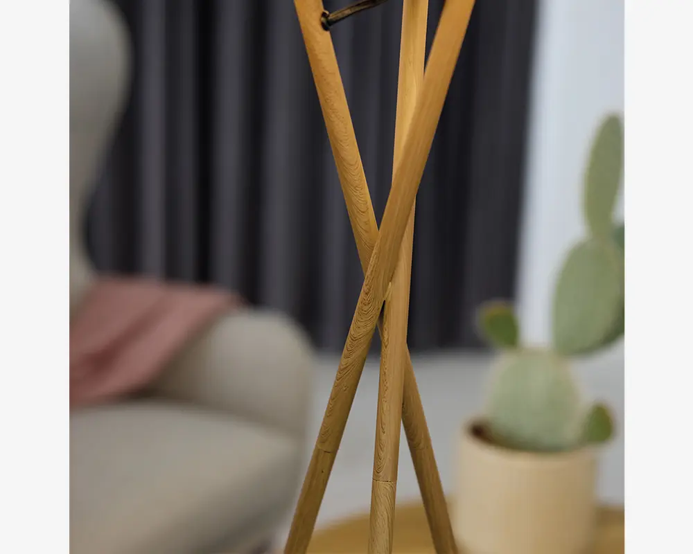 Tre slanke, lyse træstænger krydser hinanden og står oprejst, hvilket danner en tripod-struktur. Træet har en glat, naturlig finish med synlige åremønstre. Toppen af stængerne er samlet med en lille metalforbindelse. Designet er minimalistisk og moderne, velegnet til nutidige indretninger.