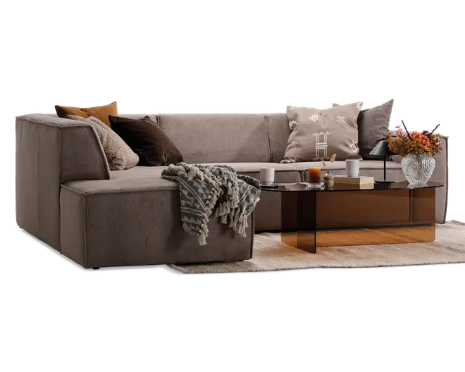 Moderne L-formet sofa betrukket med blødt, taupefarvet stof. Sofaen har brede, lavprofilerede armlæn og ryglæn og er pyntet med flere bløde pyntepuder i neutrale og jordnære nuancer som beige, brun og rust. Et tekstureret, frynset tæppe er afslappet lagt over den ene arm. Foran sofaen står et rektangulært sofabord i røgfarvet glas med dekorative genstande som stearinlys, bøger og en vase med en buket blomster i efterårsfarver. Opsætningen står på et lyst, tekstureret gulvtæppe og skaber en hyggelig og indbydende stue.