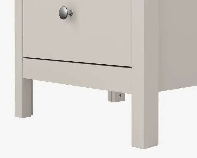 Nærbillede af den nederste del af et møbel med en glat, lysebeige overflade. Det har lige, solide ben og en skuffe med en rund, metalfarvet knop. Designet er minimalistisk og moderne med rene linjer og en lille åbning under skuffen, som gør det nemt at rengøre eller opbevare ting.