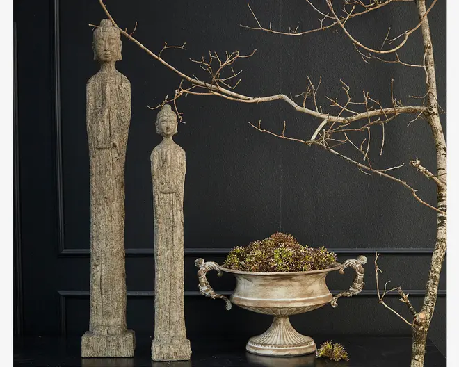 To høje, slanke dekorative statuer med rustik, patineret stenfinish står side om side. Hver statue har aflange kroppe og minimalistiske detaljer, hvor den ene er lidt højere end den anden. Ved siden af står en udsmykket, vintage-inspireret metalurne med to håndtag, fyldt med grønne og brune sukkulenter. Baggrunden består af en mørk væg og en bar, forgrenet gren, hvilket skaber et sofistikeret og naturligt udtryk.