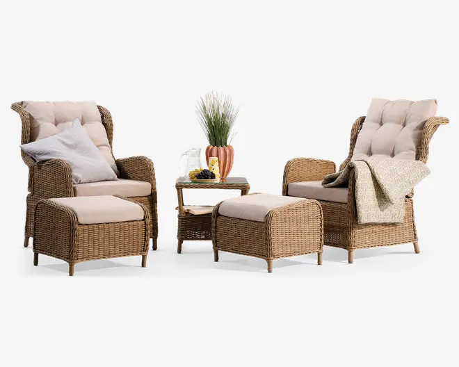 Sættet består af to høje kurvestole med tykke, beige, knap-lukkede hynder, to matchende fodskamler og et lille firkantet sidebord. Den ene stol har en hvid og en beige pude, mens den anden har et lyst, mønstret tæppe lagt over armlænet. Bordet har en dekorativ plante, to glas og en tallerken med frugt på toppen samt en bog på nederste hylde. Kurvematerialet er i en naturlig, lys brun farve, og stilen er hyggelig og indbydende, velegnet til både inde- og udendørs brug.