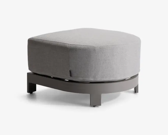 En moderne, rund puf med en lys grå stofpude og en solid, buet base i en matchende neutral farve. Puden har et glat, sømløst udseende med diskrete syninger langs kanterne, og basen har fire korte ben for stabilitet. Det samlede design er minimalistisk og moderne, velegnet til forskellige indretninger.