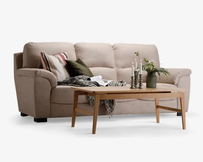 En beige tresæders sofa med bløde puder og afrundede armlæn. Sofaen er pyntet med forskellige pyntepuder i stribede, ensfarvede og teksturerede designs samt et plaid tæppe, der er lagt over. Foran sofaen står et lyst træ-sofabord med skrå ben. På bordet er der tre metal-lysestager med hvide stearinlys, en grøn vase med grønne planter og et åbent magasin, hvilket skaber en hyggelig og indbydende stemning.