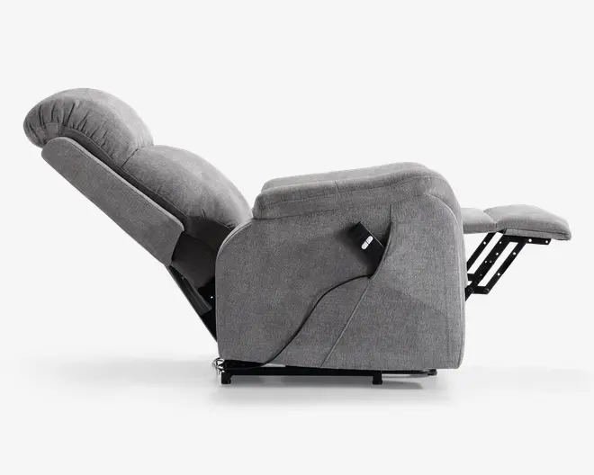 Sidebillede af en fuldt tilbagelænet stol betrukket med lysegråt stof. Stolen har en kraftigt polstret ryg, sæde og armlæn for ekstra komfort. En fastgjort fjernbetjening med ledning ses på siden, hvilket indikerer elektrisk tilbagelæningsfunktion. Fodstøtten er slået ud, og stolens bund er let hævet fra gulvet.