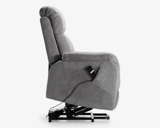 Sidebillede af en grå polstret reclinerstol med blød, polstret ryg- og armlæn. Stolen har en synlig metal løftemekanisme i bunden, som hjælper med at rejse sig op. En ledningsbaseret fjernbetjening er fastgjort på siden, hvilket indikerer justerbare indstillinger for komfort og løftefunktioner. Stoffet ser blødt og tekstureret ud, hvilket giver en hyggelig siddeoplevelse.