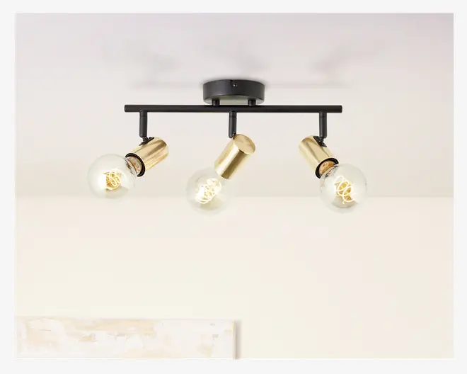 Loftmonteret lampe med tre justerbare lampehoveder. Lampen har en mat sort vandret stang og rund base, hvor hvert lampehoved er i børstet guld. Pærerne er synlige, runde og har synlige glødetråde, hvilket giver et moderne, industrielt udtryk. Hvert lampehoved kan vippes, så lyset kan rettes efter behov.